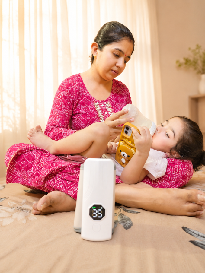 Mother feeding baby with Nestush bottle warmer beside her on bed